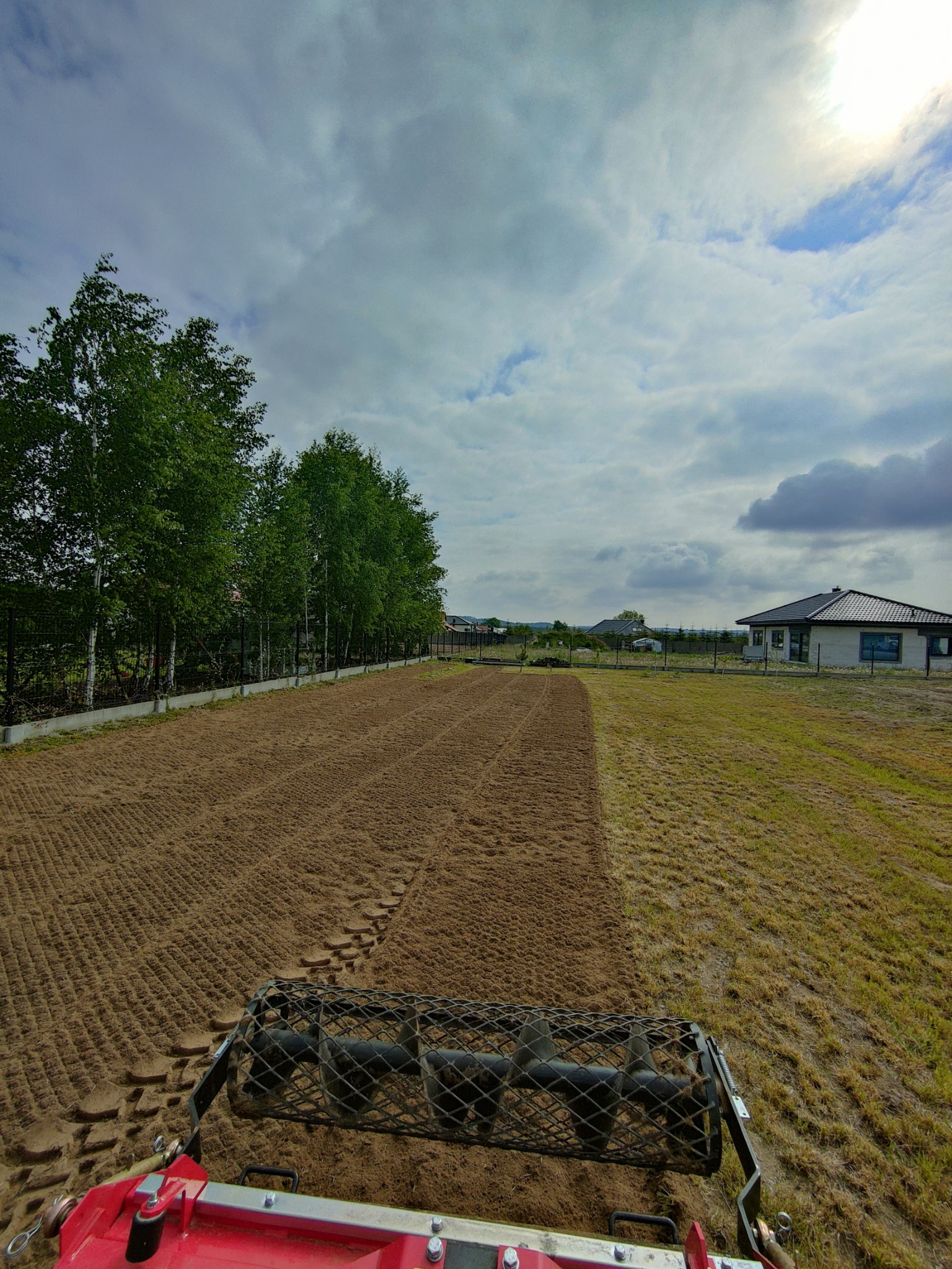 Widok z perspektywy operatora na walec glebowy ciągnięty przez traktor, przygotowujący teren pod przyszły trawnik, z widocznymi śladami pracy na wyrównanej ziemi i fragmentem domu w tle.