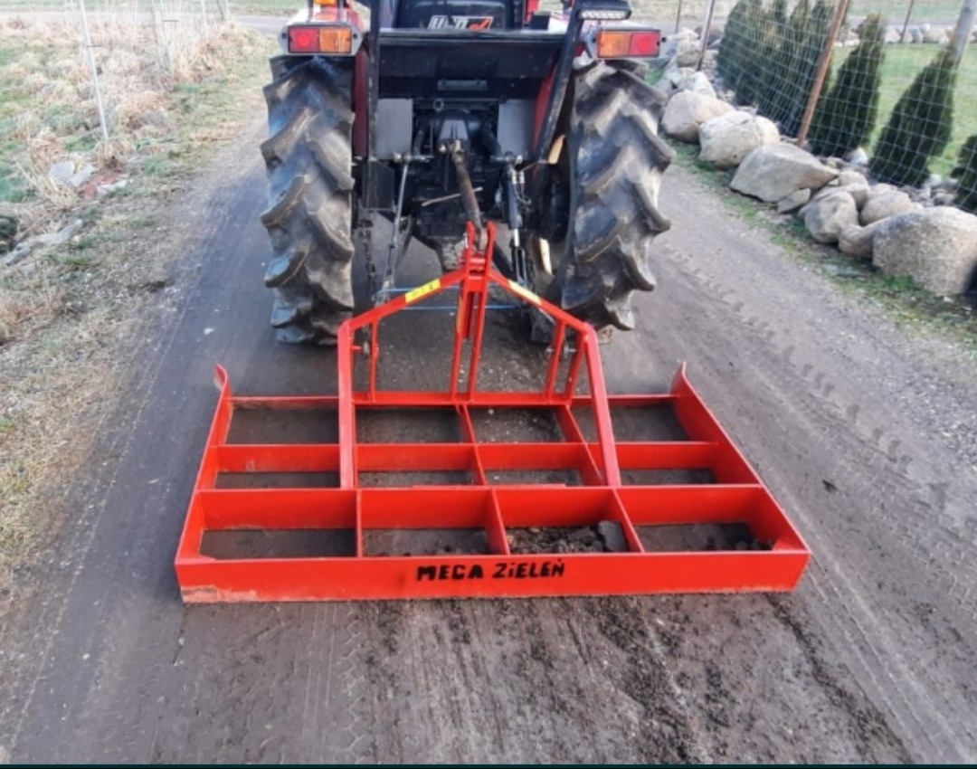 Czerwony, stalowy równiarka podczepiona do traktora na ubitej, polnej drodze. Równiarka ma kwadratową konstrukcję z wieloma przegrodami i napisem 'MEGA ZIELEN'.