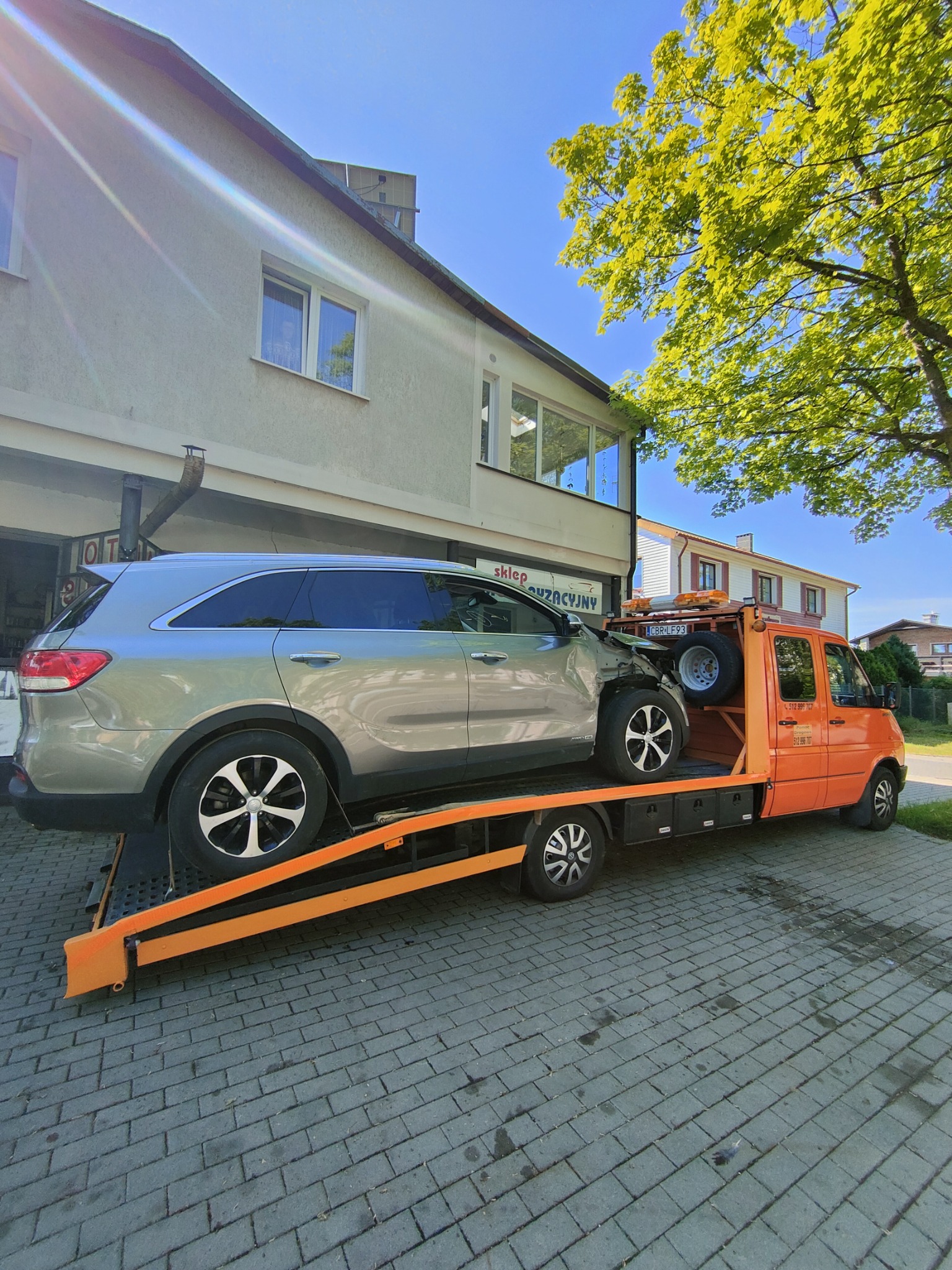 Uszkodzony srebrny SUV na pomarańczowej lawecie, zaparkowany na brukowanej ulicy przed budynkiem z jasną elewacją w słoneczny dzień.
