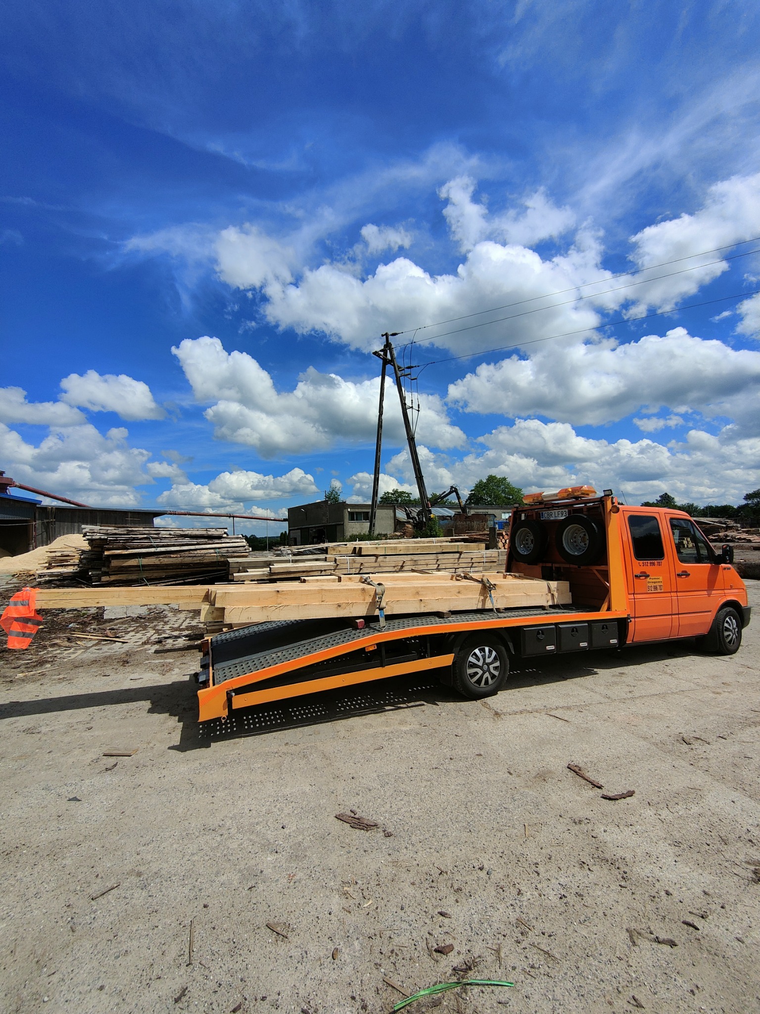 Pomarańczowa laweta z drewnem na platformie, zaparkowana na placu składowym z widocznymi deskami i budynkami w tle, błękitne niebo z obłokami.