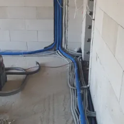 Niebieskie i szare rury karbowane z kablami elektrycznymi prowadzone wzdłuż ściany z betonu komórkowego w surowym stanie, widoczna puszka elektryczna i okno w tle.