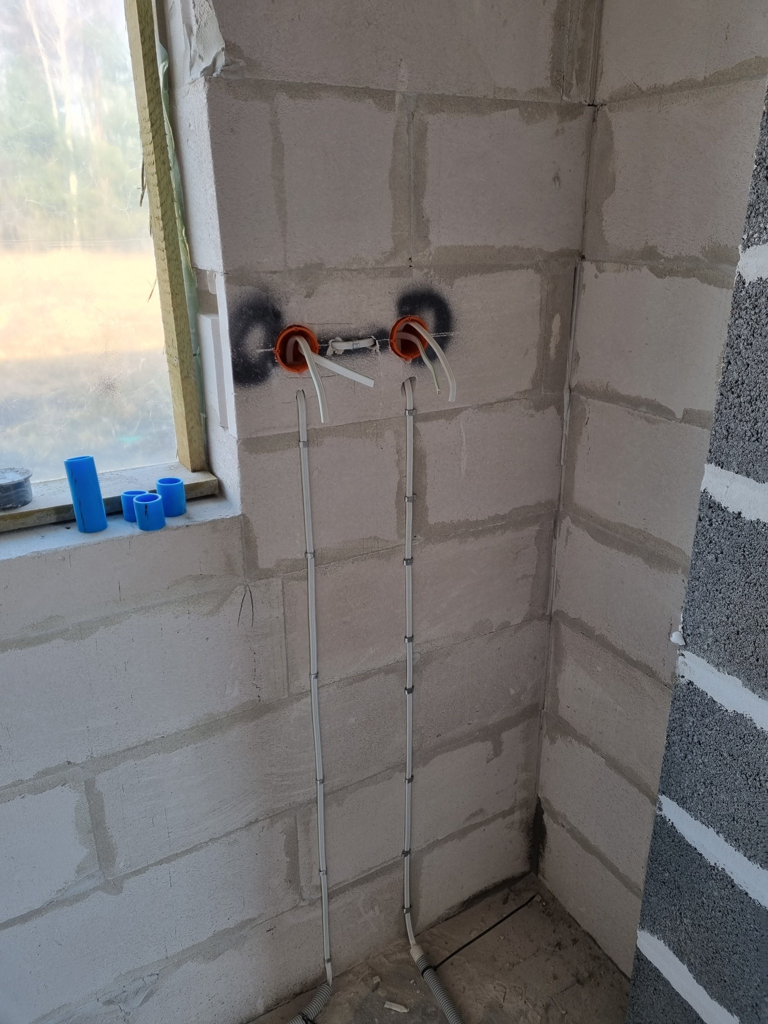 Wewnętrzna instalacja elektryczna w surowym pomieszczeniu z białych bloczków, widoczne dwa punkty wyprowadzenia przewodów w pomarańczowych puszkach, kable przytwierdzone do ściany, niebieskie rurki...