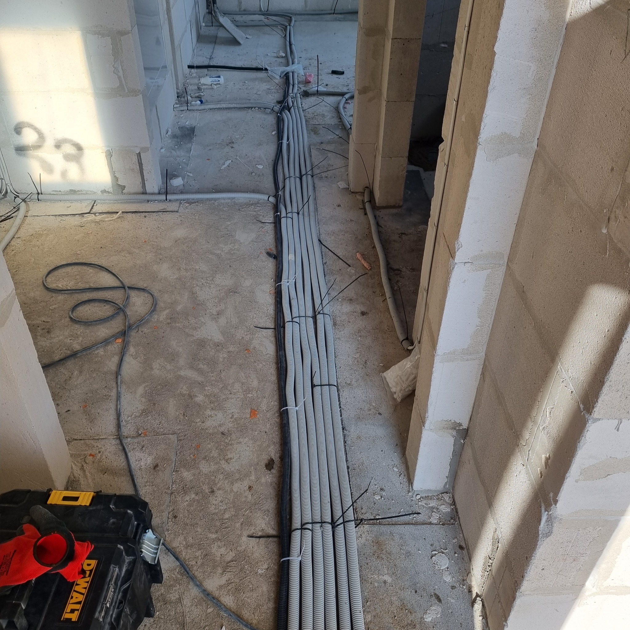 Wiązka szarych rurek karbowanych z kablami, spięta czarnymi opaskami zaciskowymi, ułożona na surowym betonowym podłożu w trakcie prac instalacyjnych w budynku, obok skrzynka narzędziowa DeWalt...