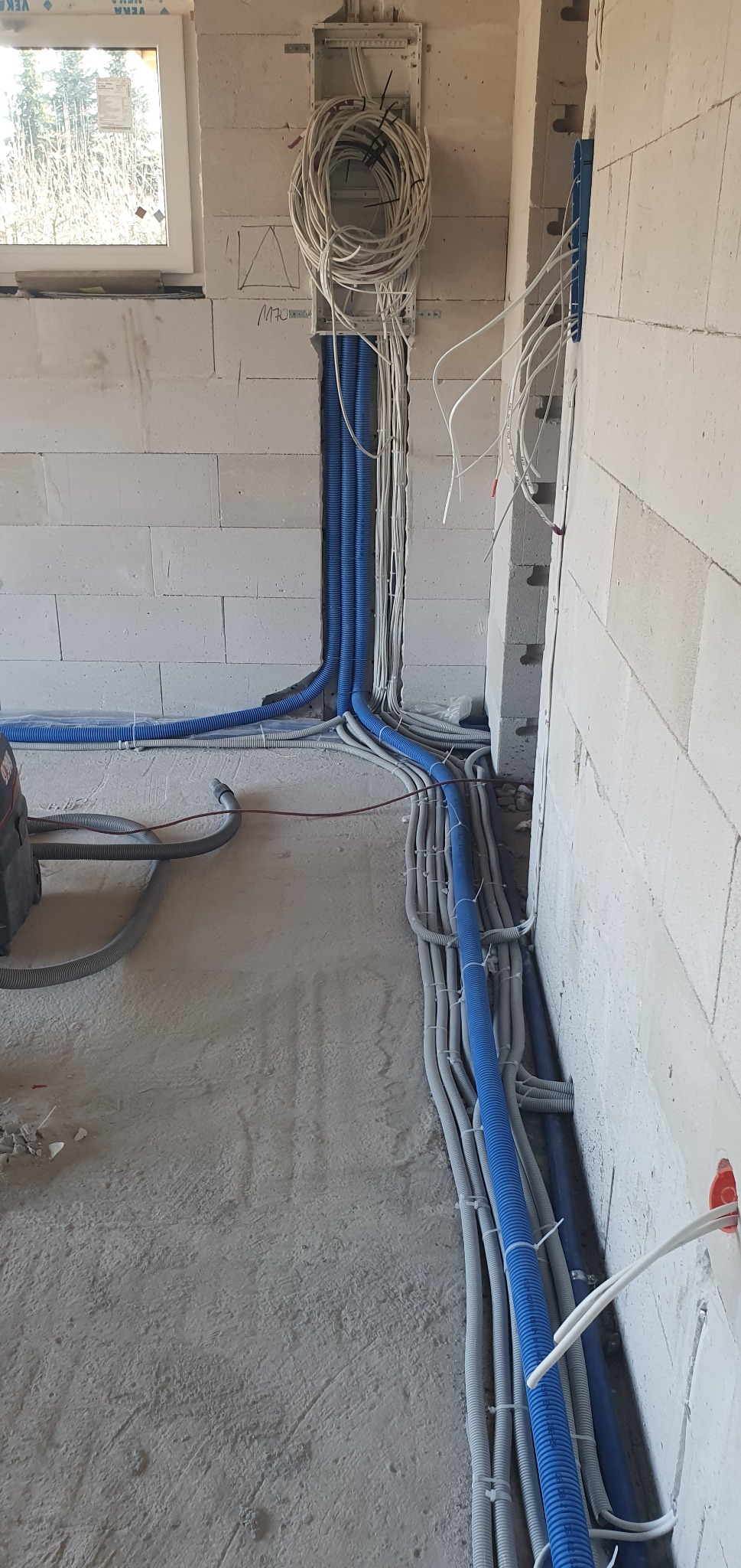 Niebieskie i szare rury karbowane z kablami elektrycznymi prowadzone wzdłuż ściany z betonu komórkowego w surowym stanie, widoczna puszka elektryczna i okno w tle.