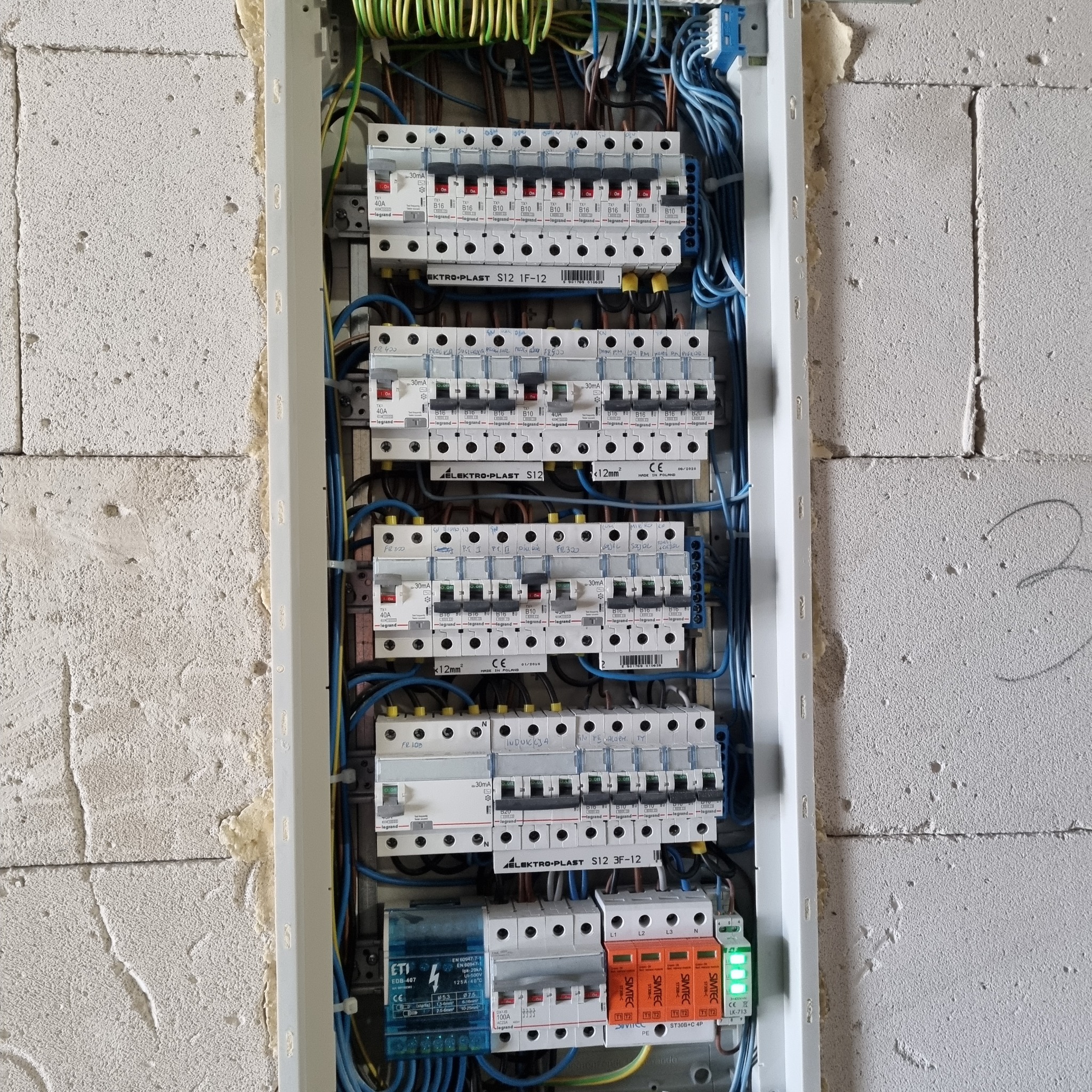 Otwarta skrzynka elektryczna z zamontowanymi bezpiecznikami i przewodami, widok na ścianę z betonu komórkowego w tle.