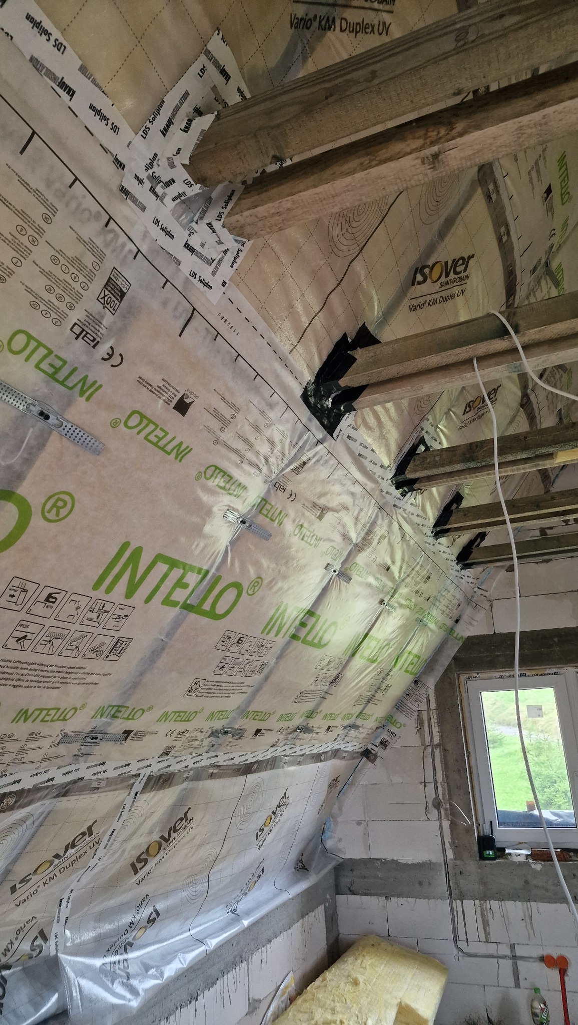 Montaż folii paroizolacyjnej Intello i Isover Vario KM Duplex UV na poddaszu, widoczne drewniane belki, okno i fragment ściany z betonu komórkowego.
