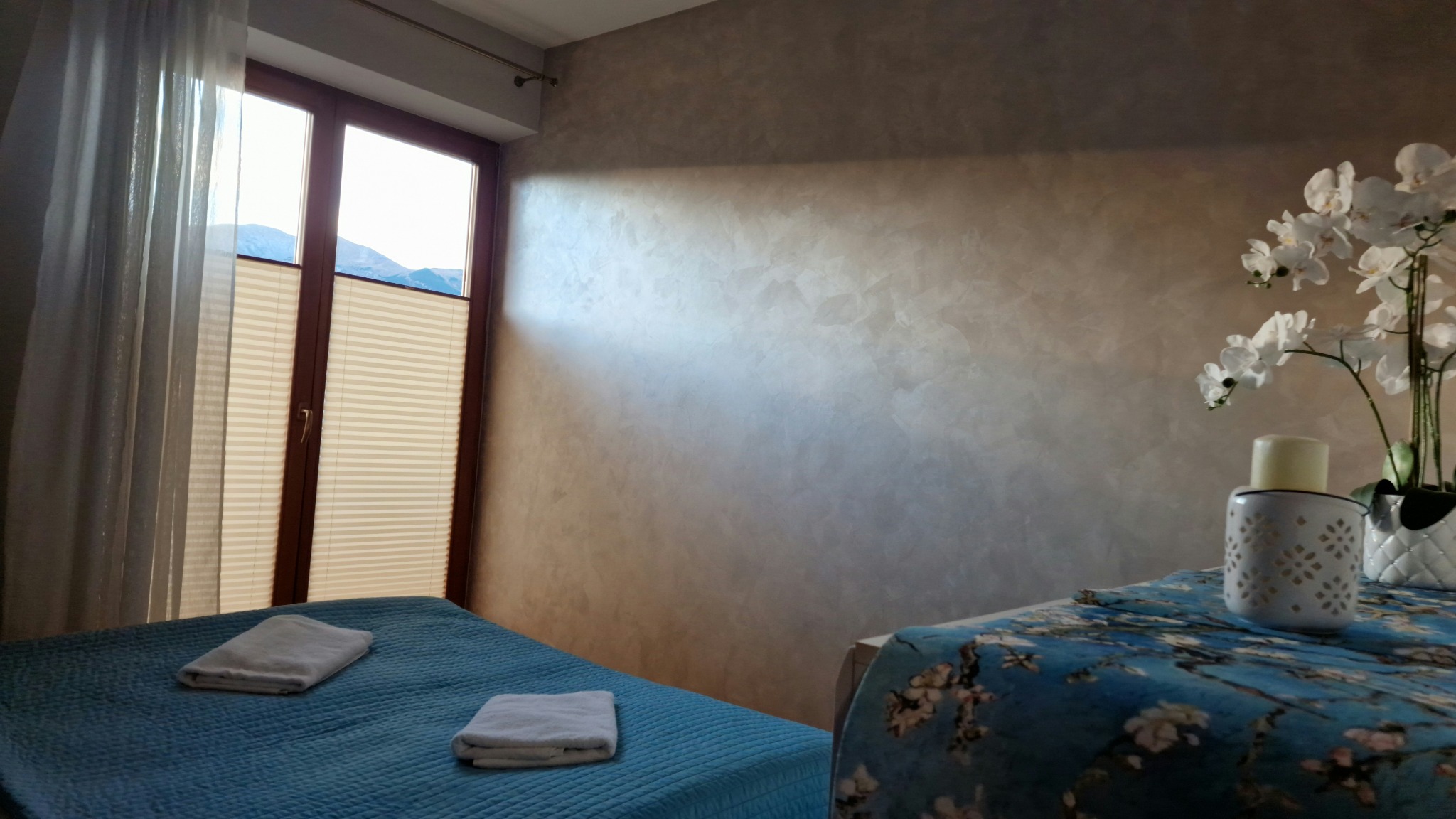 Ściana w pokoju po wykonaniu dekoracyjnego efektu tynku weneckiego, widoczne okno z żaluzjami plisowanymi i dekoracje na stoliku nocnym.