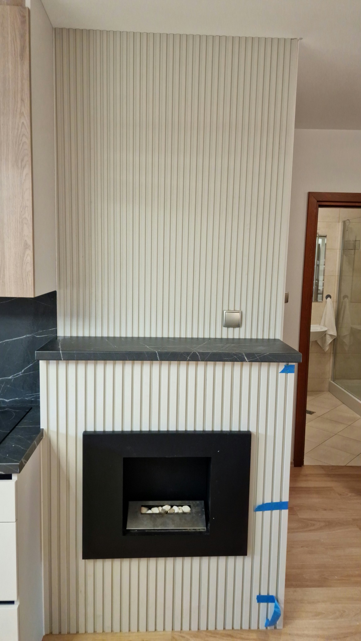 Ściana z dekoracyjnymi panelami pionowymi, z wbudowanym biokominkiem z białymi kamieniami, zwieńczona blatem z czarnego marmuru, widoczne taśmy malarskie zabezpieczające narożnik.