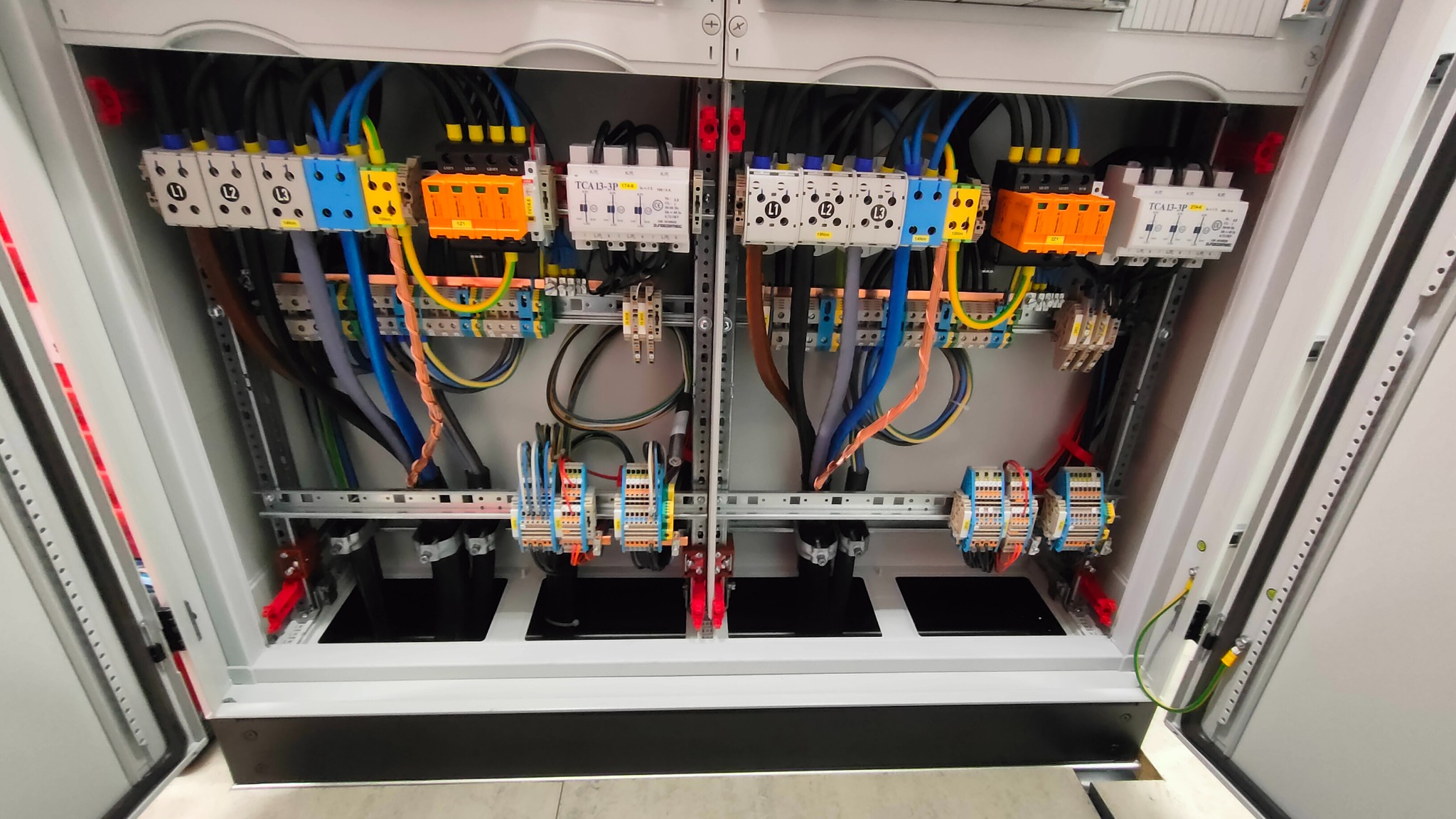 Otwarta szafa sterownicza z widocznymi przewodami, bezpiecznikami i listwami zaciskowymi, uporządkowane okablowanie w kolorach niebieskim, żółtym, brązowym i czarnym.