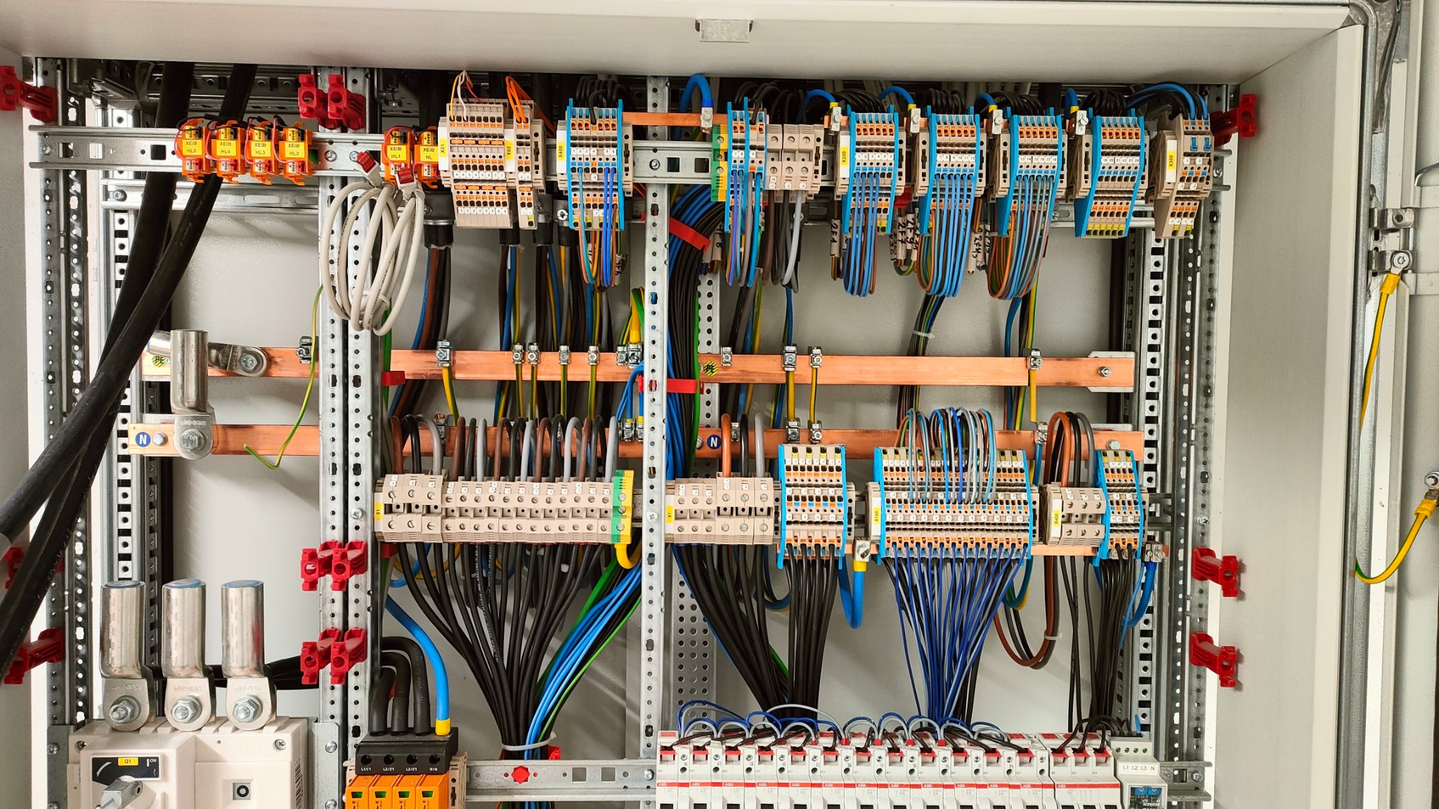 Panel elektryczny z licznymi przewodami, złączkami i zabezpieczeniami, starannie ułożonymi wewnątrz metalowej obudowy. Widoczne elementy montażowe i oznaczenia na komponentach.
