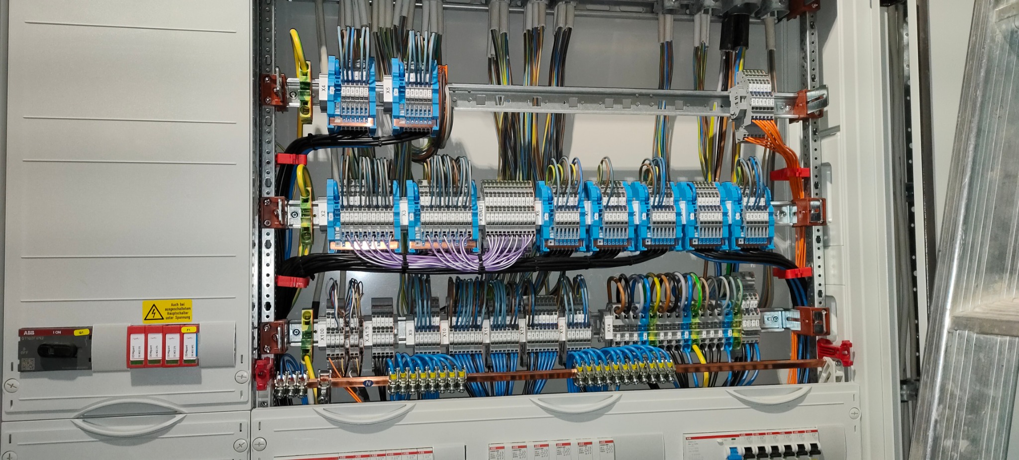 Rozdzielnica elektryczna z licznymi przewodami o różnych kolorach, zabezpieczeniami i złączkami, widoczne elementy montażowe na szynach DIN i naklejka ostrzegawcza o napięciu.