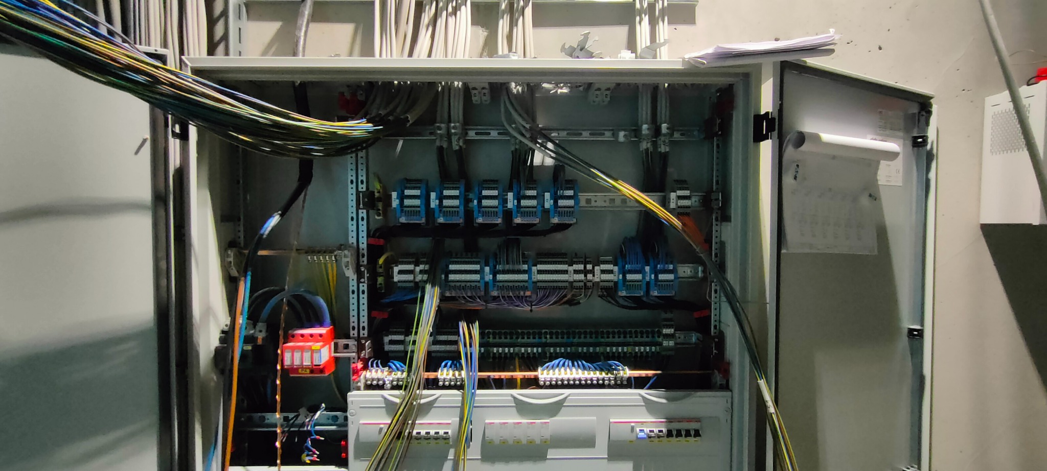 Otwarta skrzynka rozdzielcza z widocznymi bezpiecznikami, przewodami i elementami instalacji, z przewodami wychodzącymi na zewnątrz po lewej stronie, oraz dokumentacją techniczną przytwierdzoną...