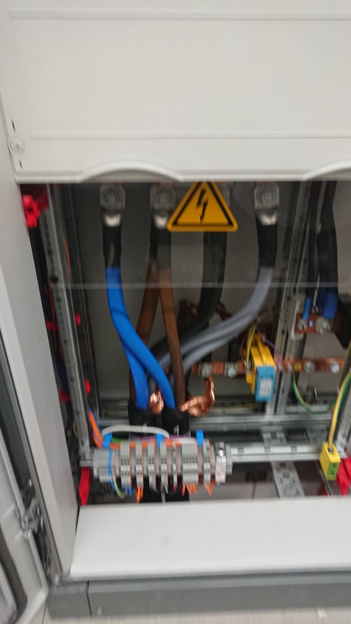 Otwarta skrzynka elektryczna z widocznymi przewodami w kolorze niebieskim, brązowym i szarym, z żółtym znakiem ostrzegawczym wysokiego napięcia, widoczne złącza i elementy montażowe.