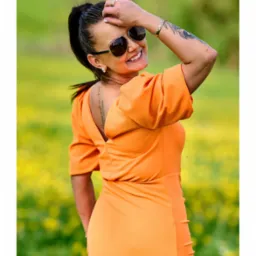 Uśmiechnięta kobieta w pomarańczowej sukience i okularach przeciwsłonecznych, z tatuażami na ramieniu i plecach, pozuje na tle kwitnącej łąki.