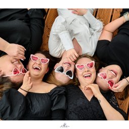 Foto Aria - Aneta Paraszkiewicz - Pięć uśmiechniętych kobiet leżących głowami do środka na drewnianym podłożu, ubrane w czarne i białe stroje, noszące okulary w kształcie serc, z widocznymi detalami jak aparat ortodontyczny i tatuaż.