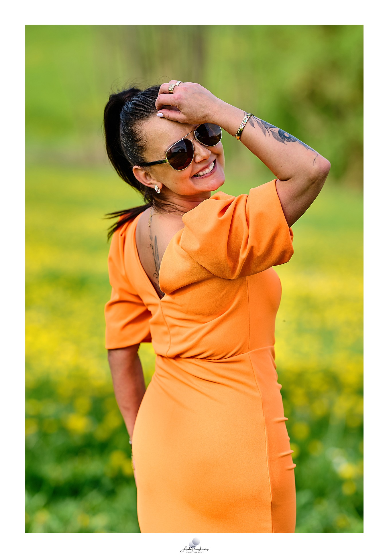 Uśmiechnięta kobieta w pomarańczowej sukience i okularach przeciwsłonecznych, z tatuażami na ramieniu i plecach, pozuje na tle kwitnącej łąki.