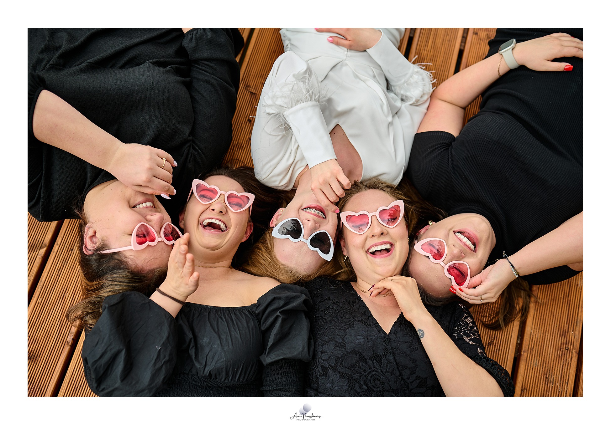 Pięć uśmiechniętych kobiet leżących głowami do środka na drewnianym podłożu, ubrane w czarne i białe stroje, noszące okulary w kształcie serc, z widocznymi detalami jak aparat ortodontyczny i tatuaż.