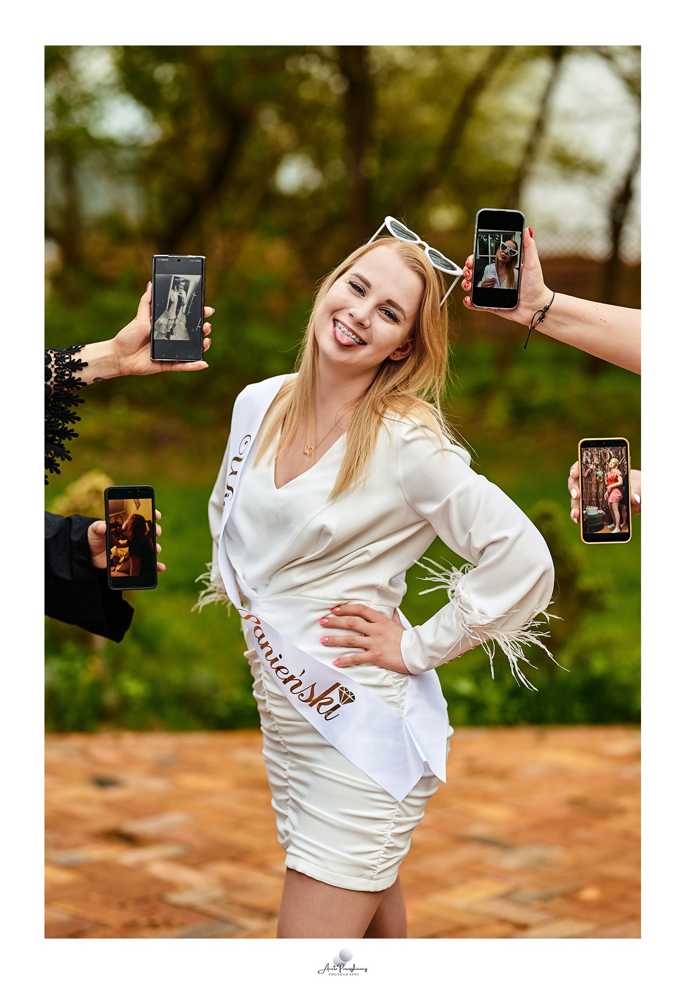 Uśmiechnięta panna młoda w białej sukience i szarfie 'Panienski' pozuje otoczona rękami trzymającymi telefony z jej zdjęciami z różnych etapów wieczoru panieńskiego.