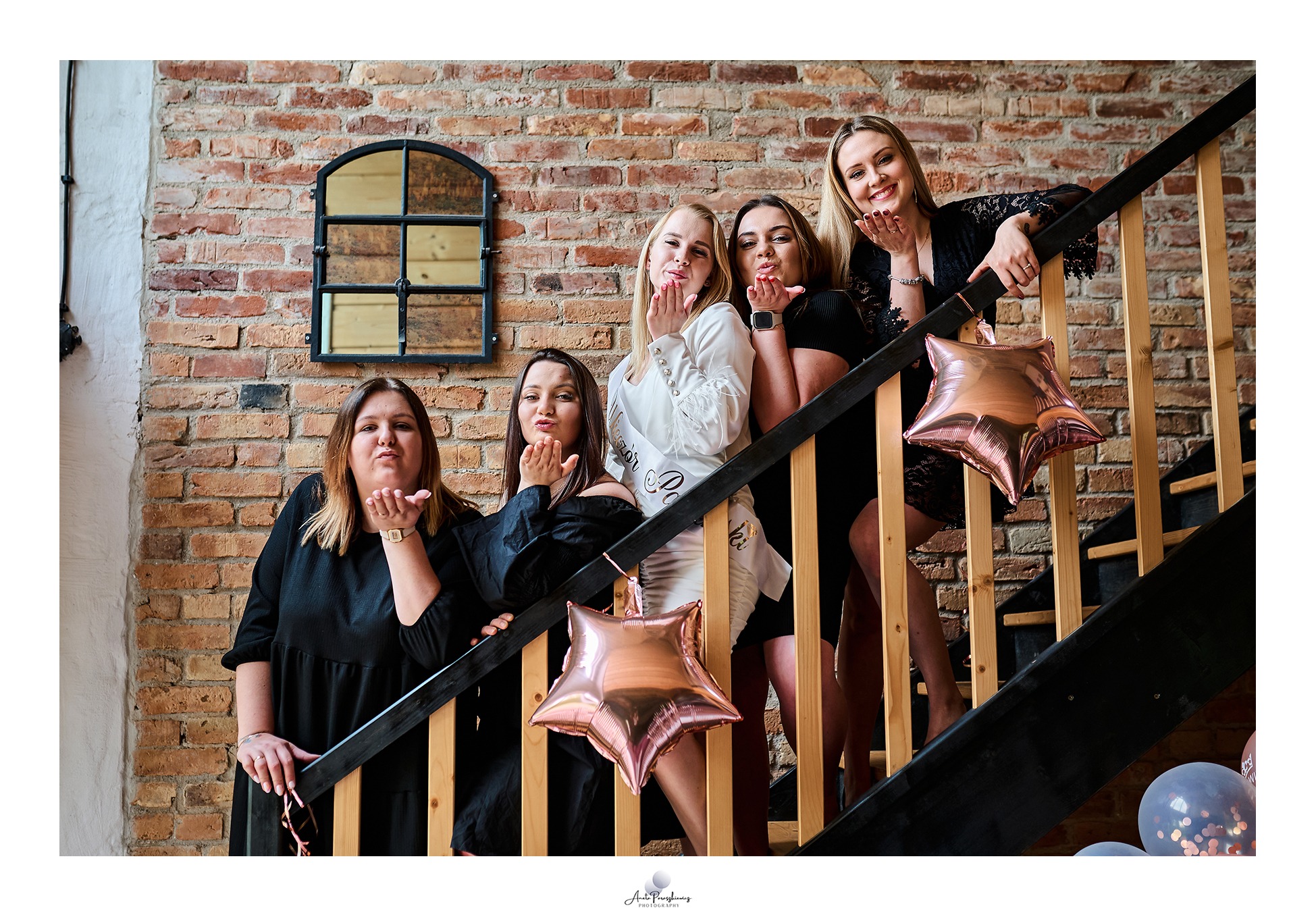 Pięć uśmiechniętych kobiet w różnym wieku, w tym jedna z szarfą 'Wieczór Panieński', pozuje na schodach z ceglaną ścianą w tle, wysyłając całusy w stronę obiektywu, z dekoracjami w postaci balonów...