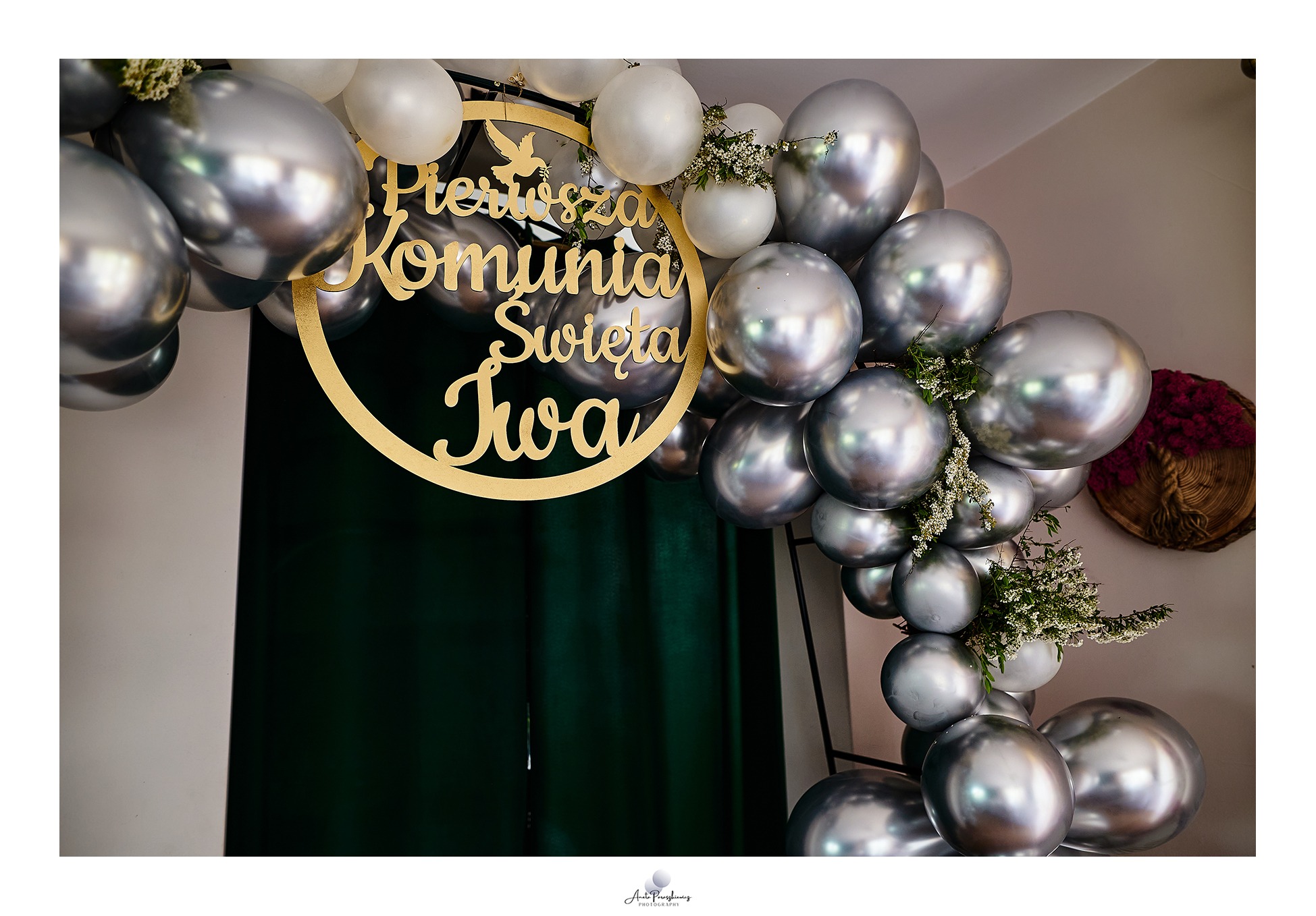 Dekoracja z srebrnych i białych balonów, złotego napisu 'Pierwsza Komunia Święta Iwa' oraz delikatnych kwiatów, tworząca łuk na tle ciemnozielonej zasłony.