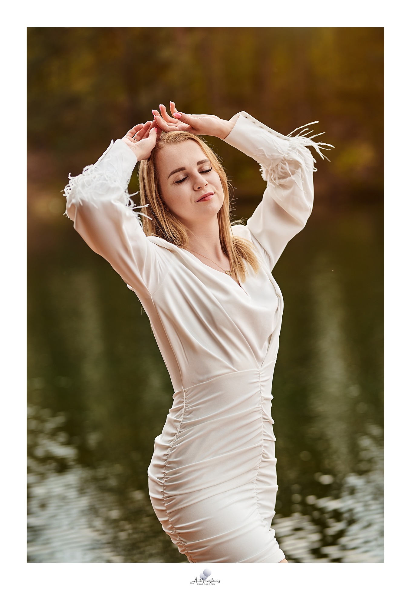 Portret kobiety w białej sukience z piórami na rękawach, pozującej z zamkniętymi oczami na tle jeziora i jesiennych drzew.
