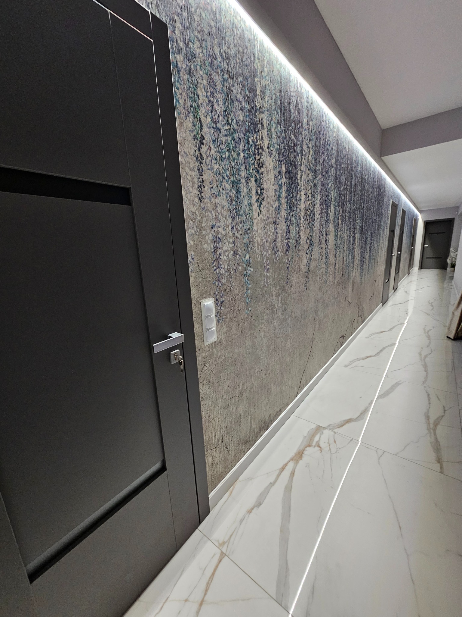 Długi korytarz z marmurową podłogą, tapetą w motywy roślinne i oświetleniem LED podsufitowym, widoczne czarne drzwi z kluczem w zamku.