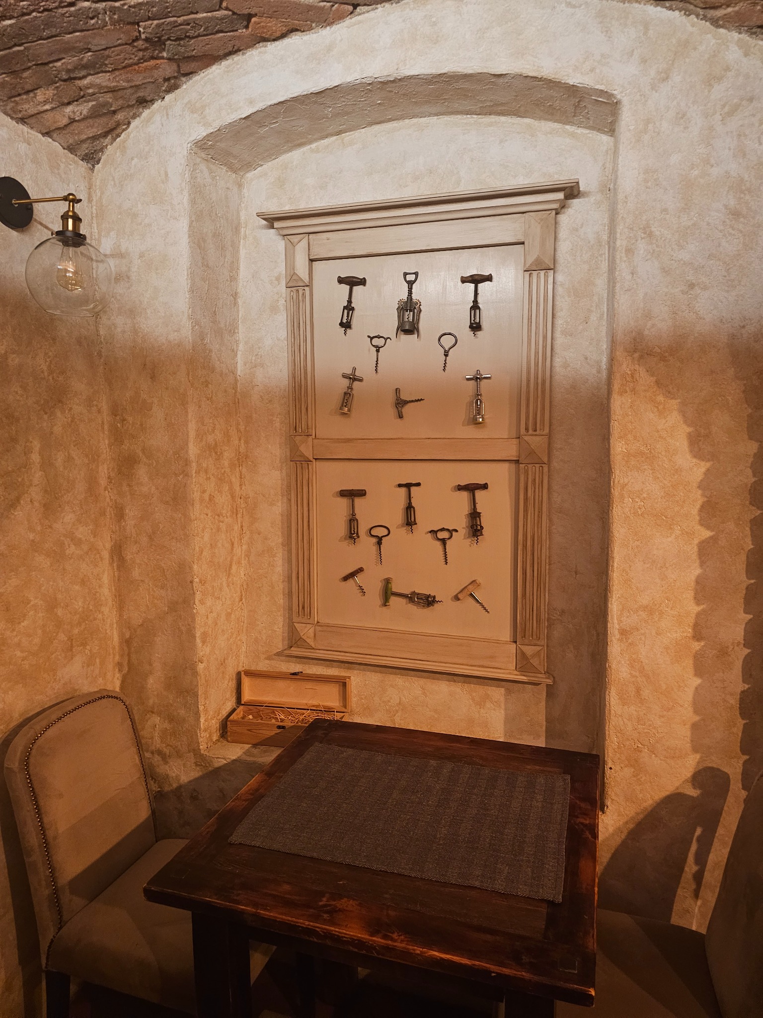 Kącik degustacyjny w piwniczce: drewniany stół z szarą podkładką, krzesło obite beżowym materiałem, ściana z wnęką eksponującą kolekcję korkociągów, oświetlona lampą z żarówką Edisona.