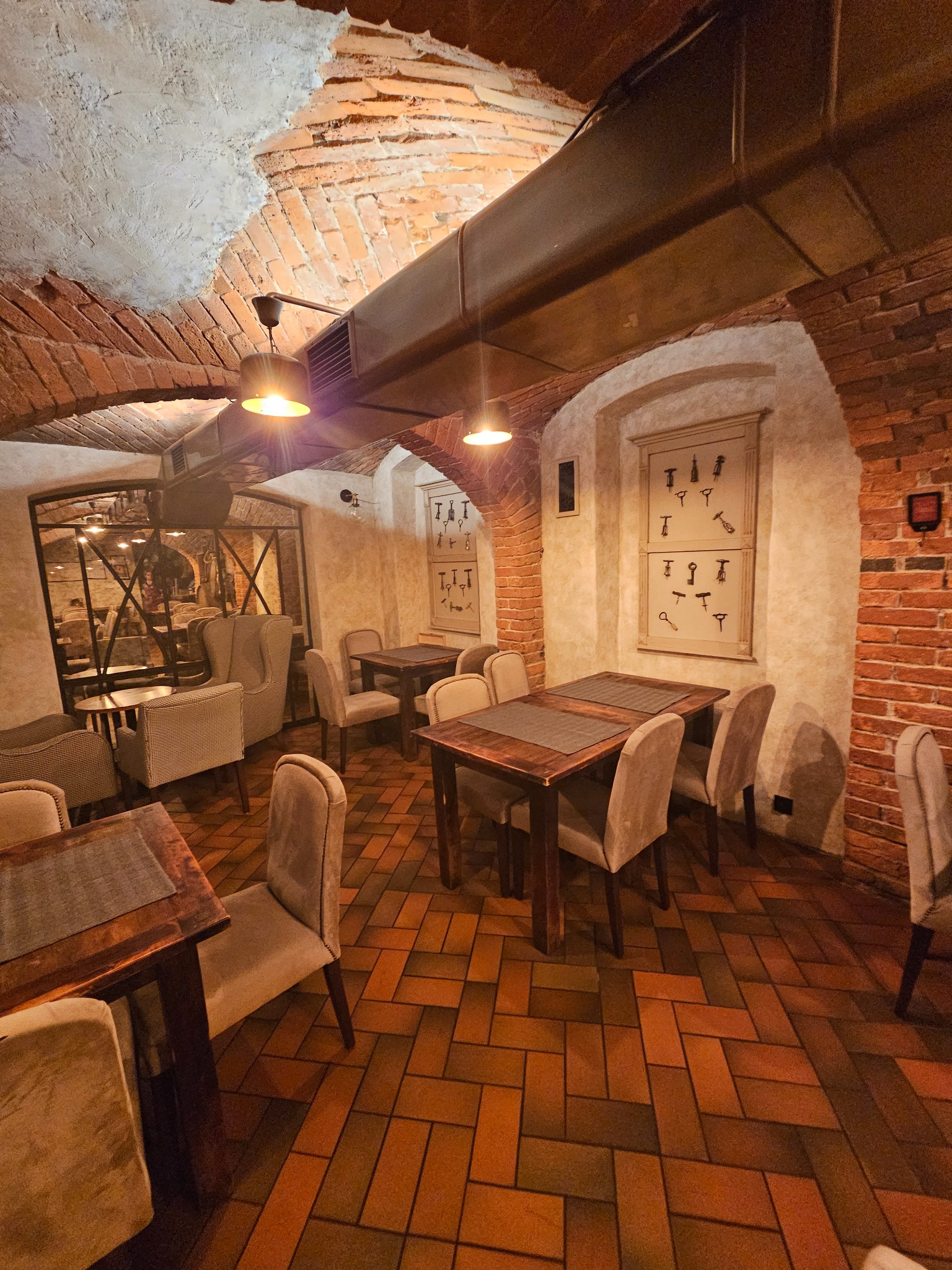 Klimatyczne wnętrze restauracji z ceglanymi łukami, drewnianymi stołami i beżowymi krzesłami, oświetlone wiszącymi lampami i z widoczną instalacją wentylacyjną pod sklepieniem.