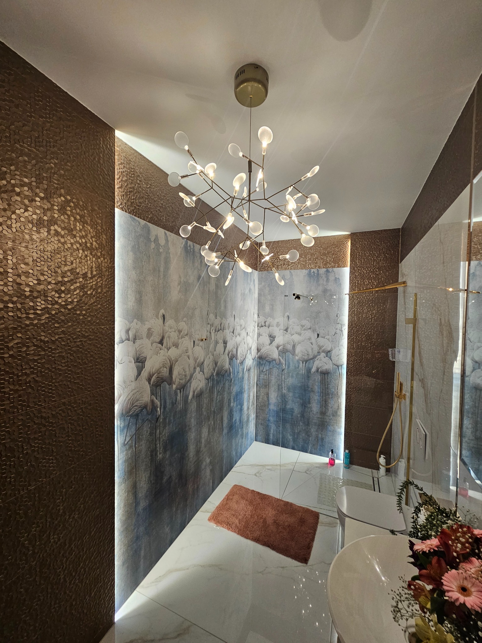 Nowoczesna łazienka z białymi marmurowymi płytkami, ścianą z motywem flamingów i designerskim, rozłożystym żyrandolem z kulistymi lampkami. Brązowe, teksturowane płytki na ścianie tworzą kontrast.