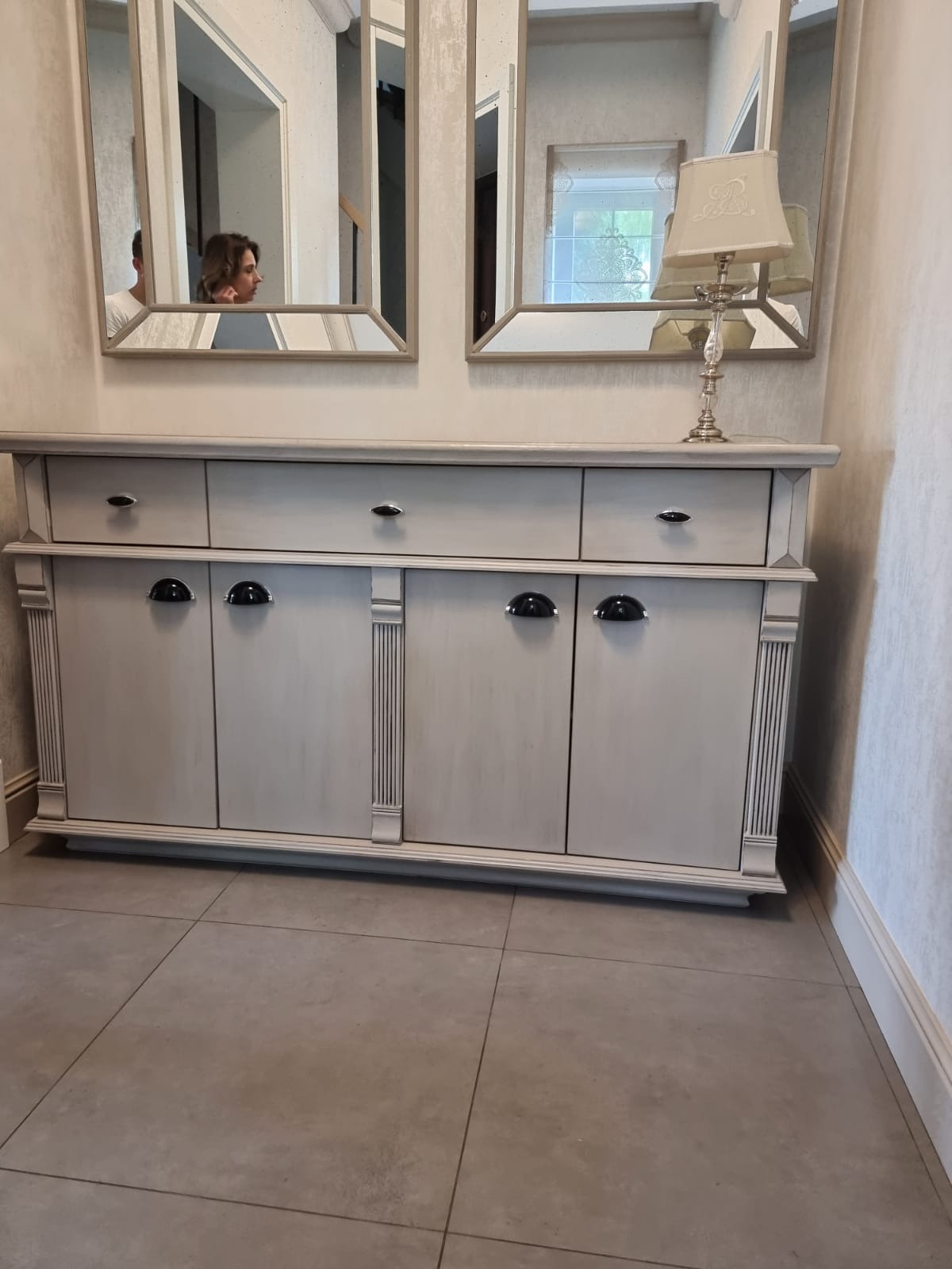 Szara komoda z trzema szufladami i trzema szafkami, nad nią dwa lustra w ozdobnych ramach i lampka nocna z abażurem, widoczna osoba w odbiciu lustra.