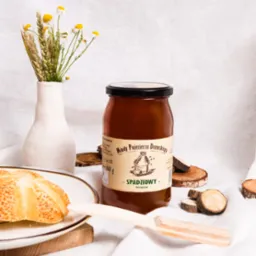 Słoik miodu spadziowego z Pojezierza Drawskiego na białym płótnie, obok wazon z polnymi kwiatami, kromka bułki z sezamem i drewniana łyżka do miodu, otoczony dekoracyjnymi plasterkami drewna.