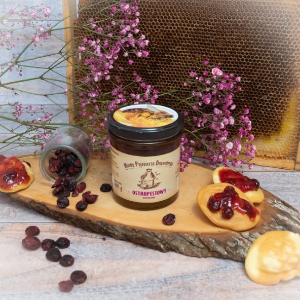Słoik miodu ostropestowego z etykietą 'Miody Pojezierza Drawskiego' na drewnianej desce, obok odwrócony słoik z suszoną żurawiną, wafle z żurawiną, plaster miodu w tle i gałązki różowych kwiatów.
