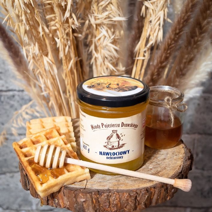 Słoik miodu nawłociowego z etykietą 'Miody Pojezierza Drawskiego' obok małego słoiczka, drewnianego nabieraka i kilku gofrów na drewnianym plastrze.