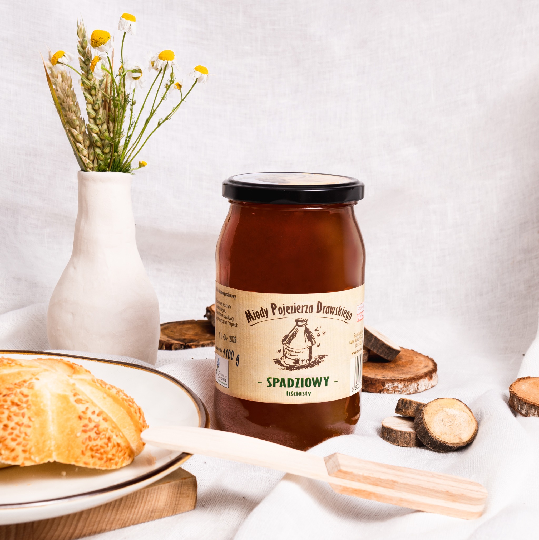 Słoik miodu spadziowego z Pojezierza Drawskiego na białym płótnie, obok wazon z polnymi kwiatami, kromka bułki z sezamem i drewniana łyżka do miodu, otoczony dekoracyjnymi plasterkami drewna.