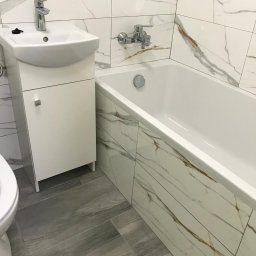 Bartłomiej Kundt - Nowoczesna łazienka z białymi płytkami imitującymi marmur, szarą podłogą imitującą drewno, białą szafką pod umywalką oraz białą wanną z zabudową.