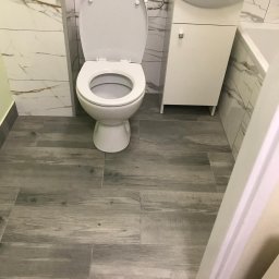 Bartłomiej Kundt - Biała toaleta z otwartą klapą, umywalka z szafką i marmurowe płytki ścienne w wyremontowanej łazience, podłoga wyłożona panelami imitującymi drewno.