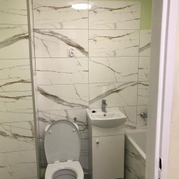 Bartłomiej Kundt - Wykończona łazienka z białymi płytkami imitującymi marmur, wisząca szafka z umywalką i toaleta z otwartą klapą.