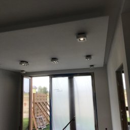 Bartłomiej Kundt - Wnętrze w trakcie remontu: sufit z zamontowanym oświetleniem punktowym, okna zabezpieczone folią malarską, widoczne ślady prac wykończeniowych na ścianach i podłodze.