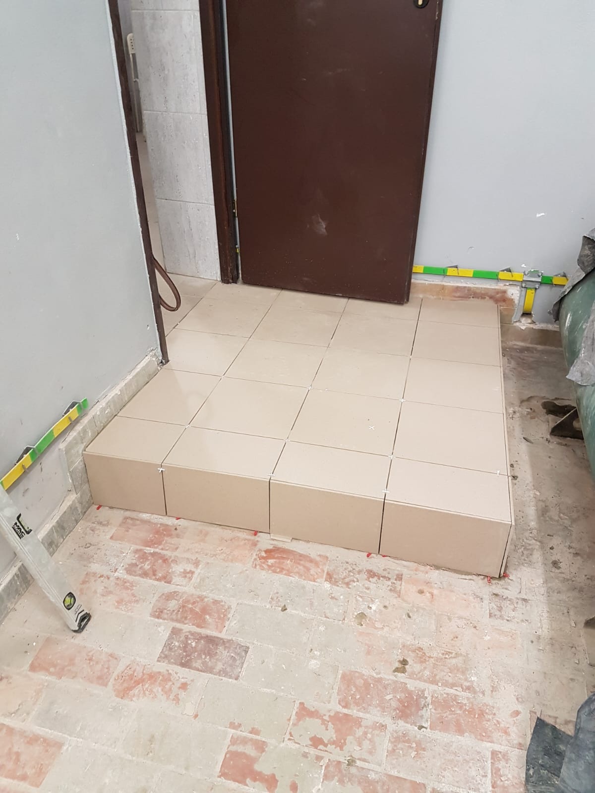 Wyrównany fragment podłogi z beżowych płytek, widoczne krzyżyki dystansowe, w tle drzwi i ściana, na podłodze stare płytki imitujące cegłę.