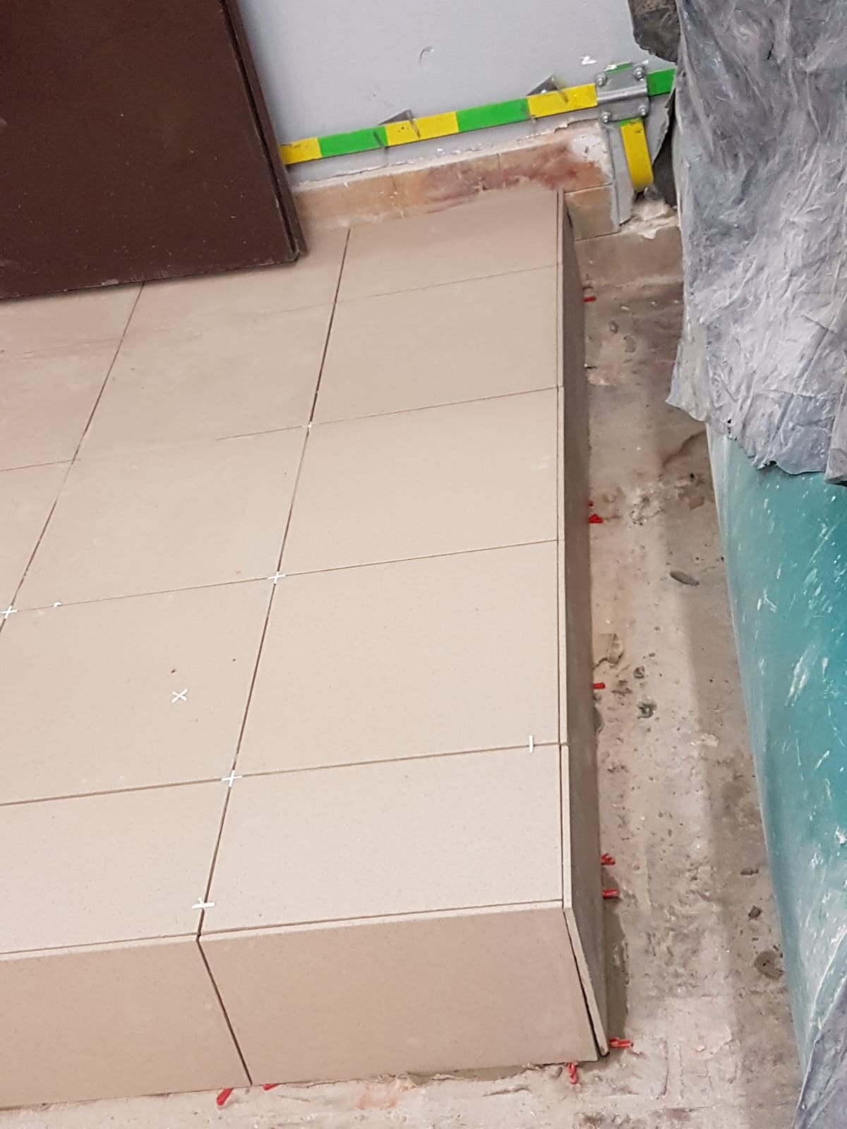 Układanie beżowych płytek podłogowych z krzyżykami dystansowymi, widoczne fragmenty podłoża i zabezpieczenia rur.