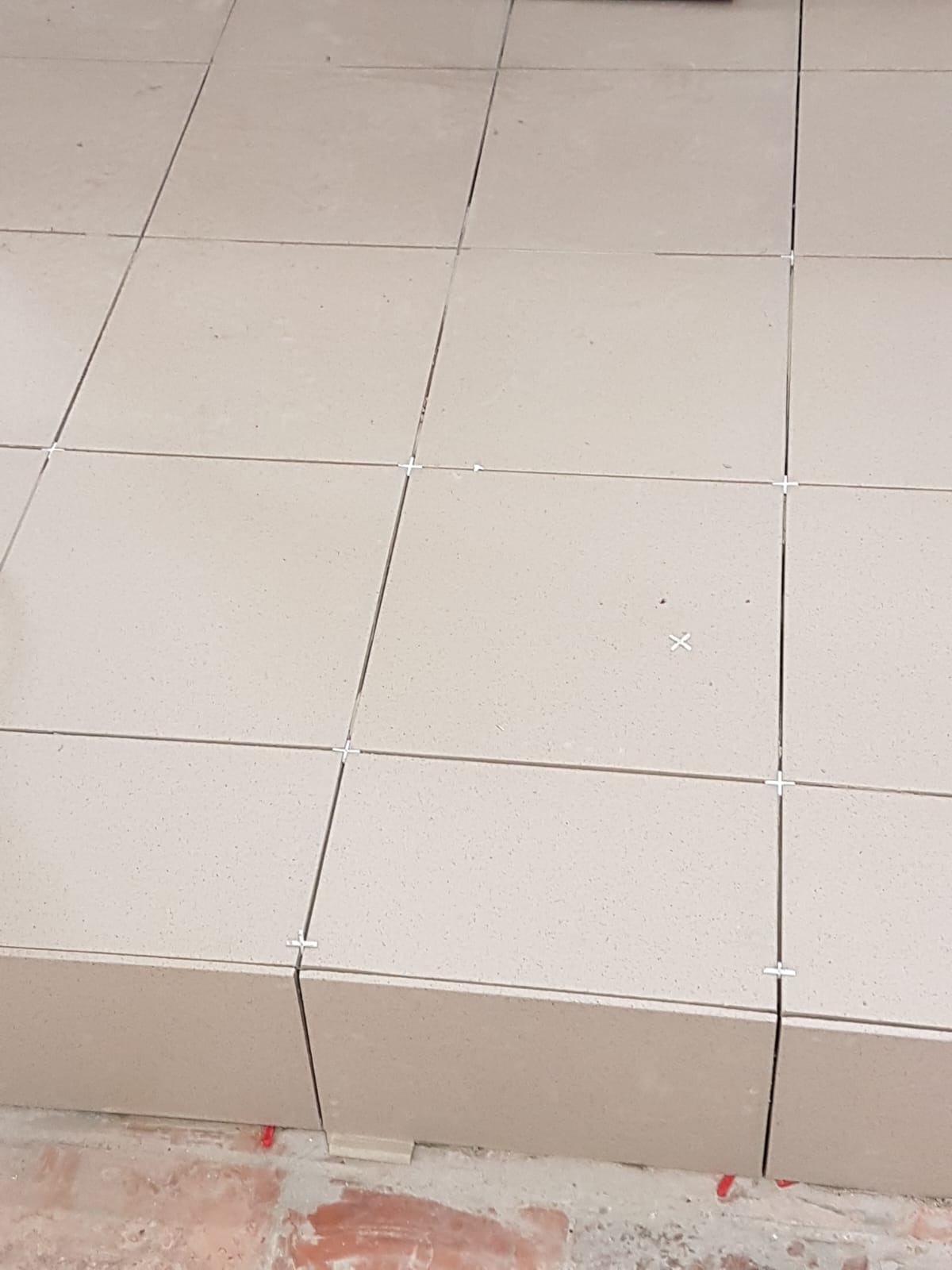 Jasne, kwadratowe płytki podłogowe ułożone z krzyżykami dystansowymi, widoczny fragment starej, ceglanej posadzki w tle.