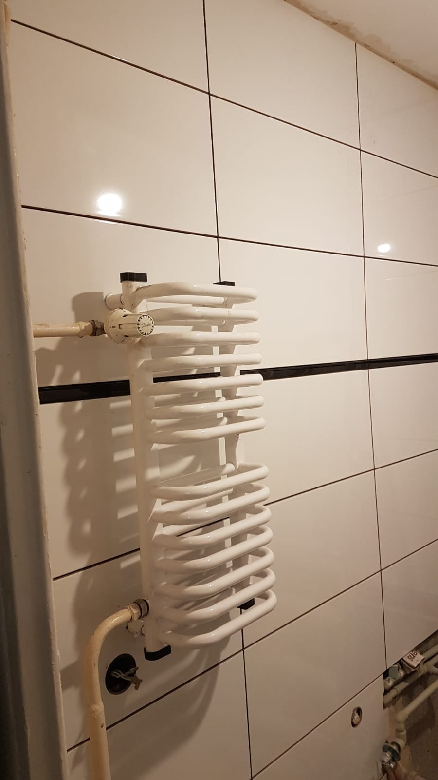 Biały, spiralny grzejnik łazienkowy na ścianie z białych płytek z ciemnymi fugami, widoczne rury doprowadzające wodę, brak wykończenia przy podłodze.