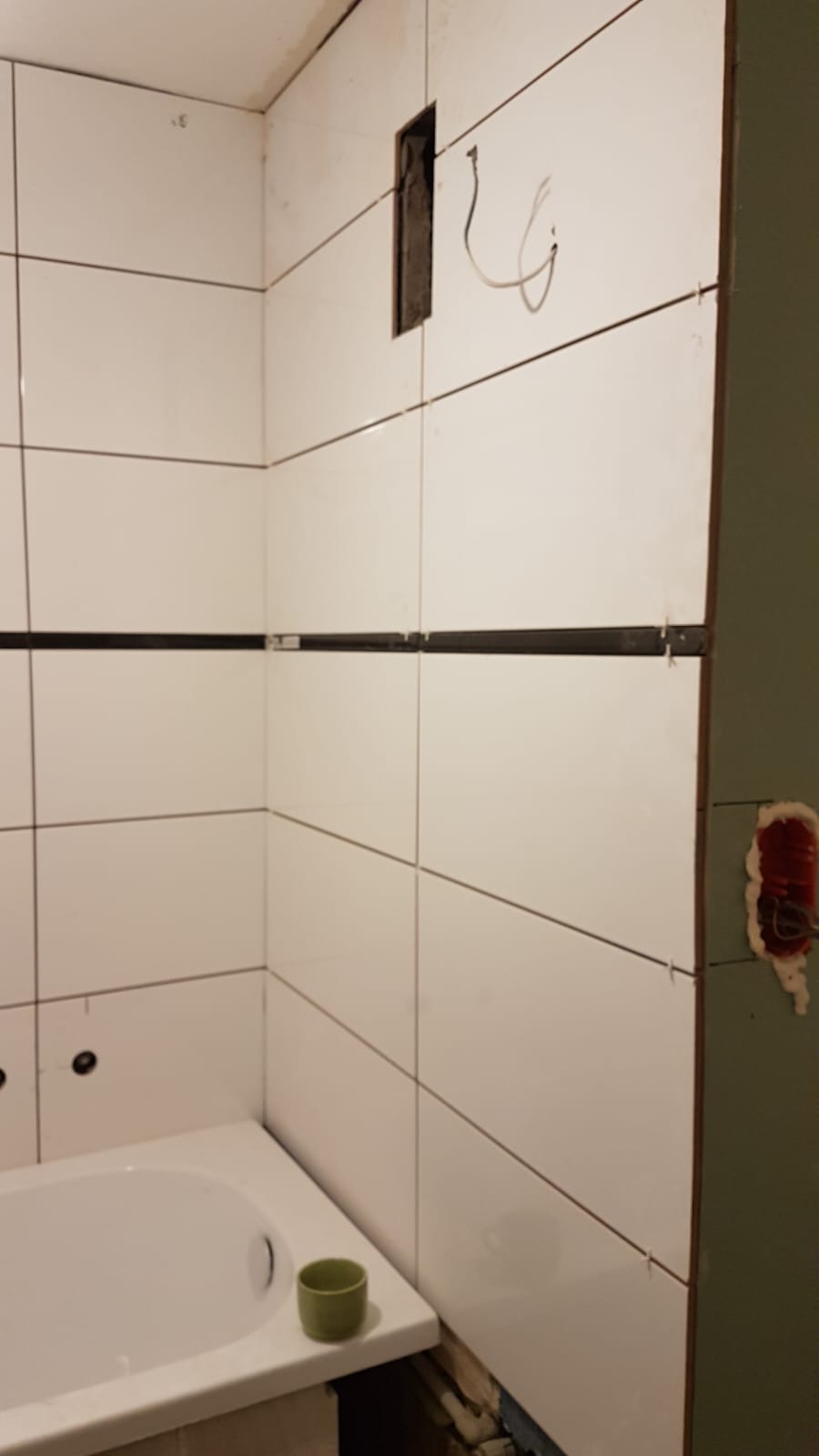 Białe płytki z czarnym paskiem w trakcie układania na ścianie w łazience, widoczny otwór na gniazdko elektryczne i wystające przewody, narożnik wanny, mały zielony kubek.