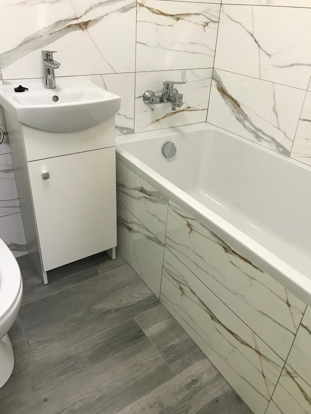 Nowoczesna łazienka z białymi płytkami imitującymi marmur, szarą podłogą imitującą drewno, białą szafką pod umywalką oraz białą wanną z zabudową.