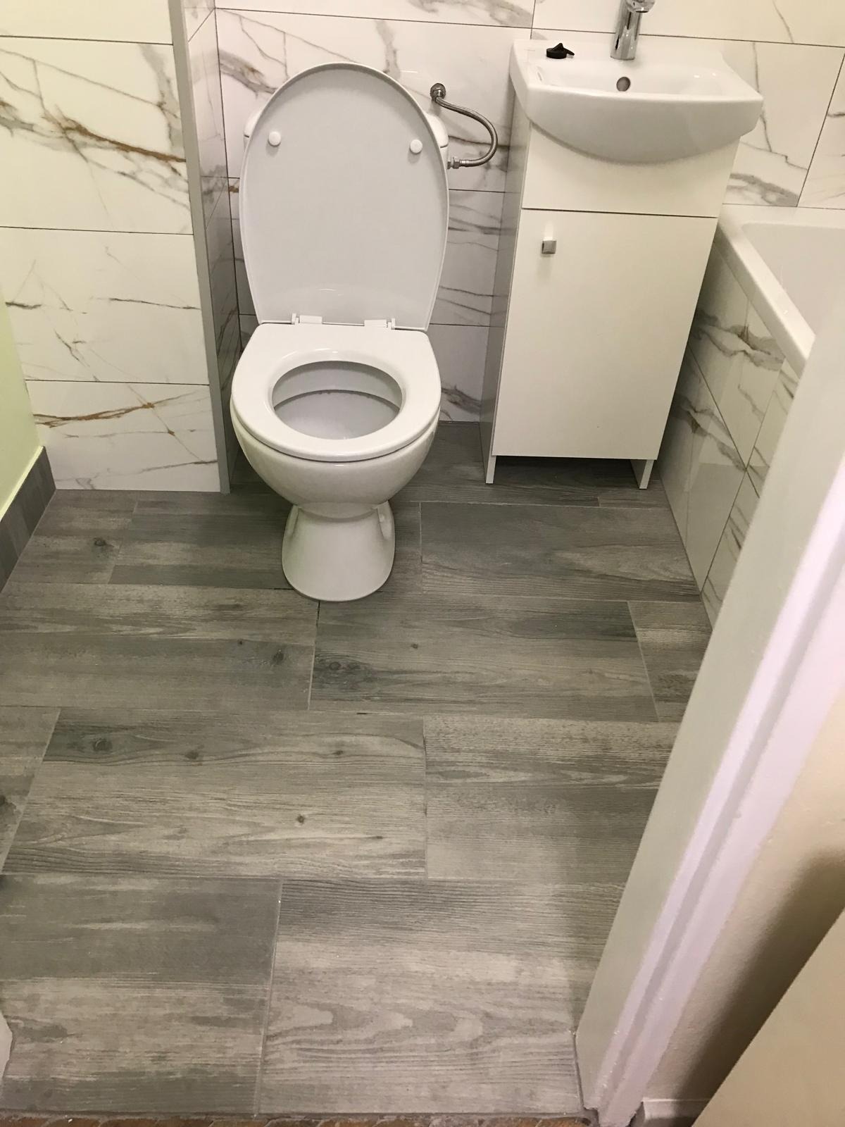 Biała toaleta z otwartą klapą, umywalka z szafką i marmurowe płytki ścienne w wyremontowanej łazience, podłoga wyłożona panelami imitującymi drewno.