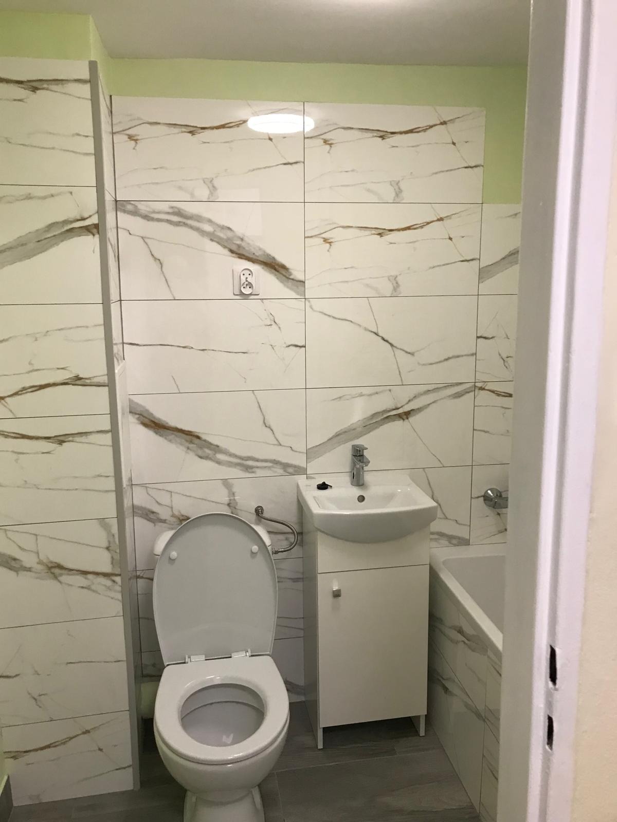 Wykończona łazienka z białymi płytkami imitującymi marmur, wisząca szafka z umywalką i toaleta z otwartą klapą.