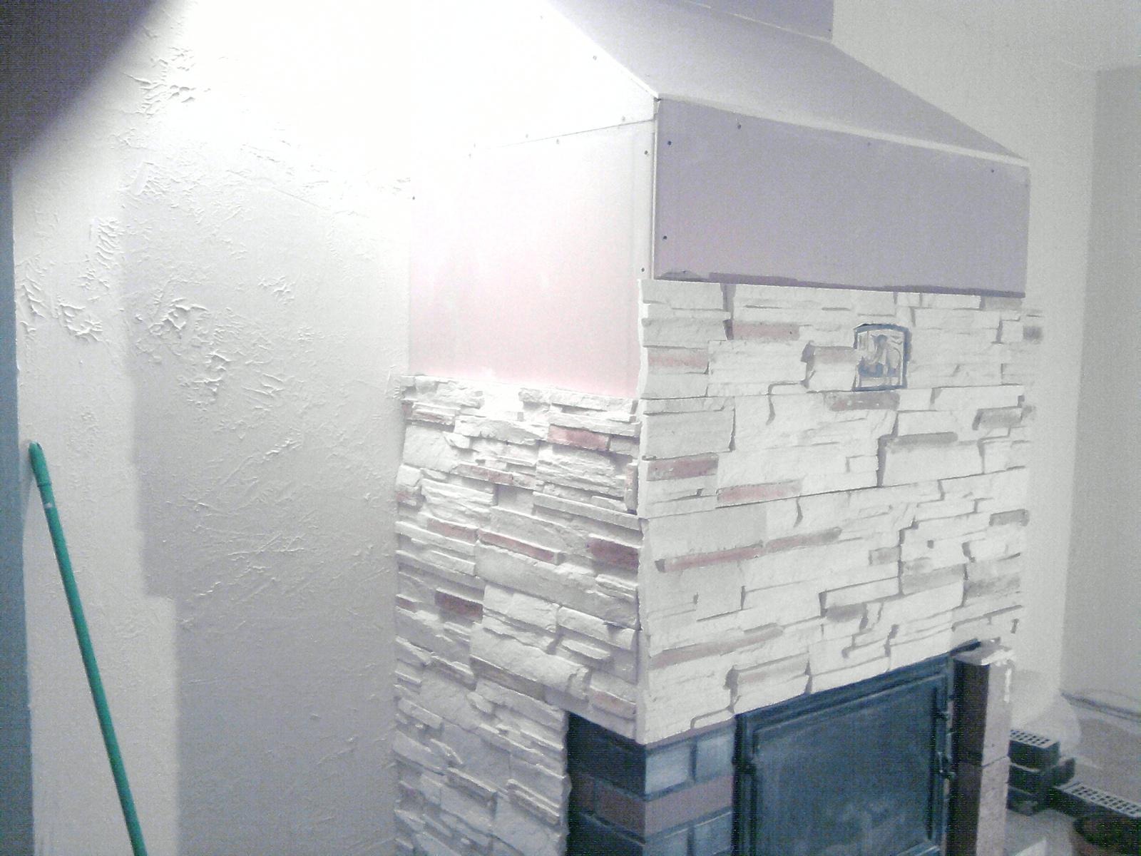 Fragment ściany z obudową kominka w trakcie wykańczania: dolna część obłożona dekoracyjnym kamieniem w odcieniach bieli i beżu, górna część w surowym stanie z płyt kartonowo-gipsowych, ściana obok...