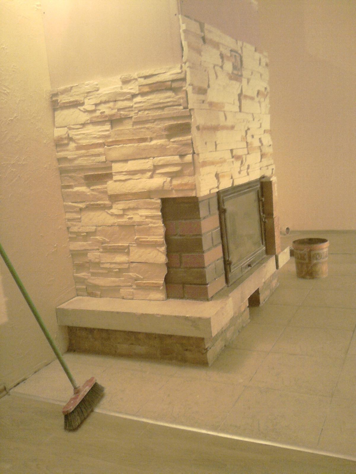 Niedokończony kominek obłożony płytkami imitującymi kamień, z widoczną metalową wkładką kominkową i szczotką do zamiatania na pierwszym planie.