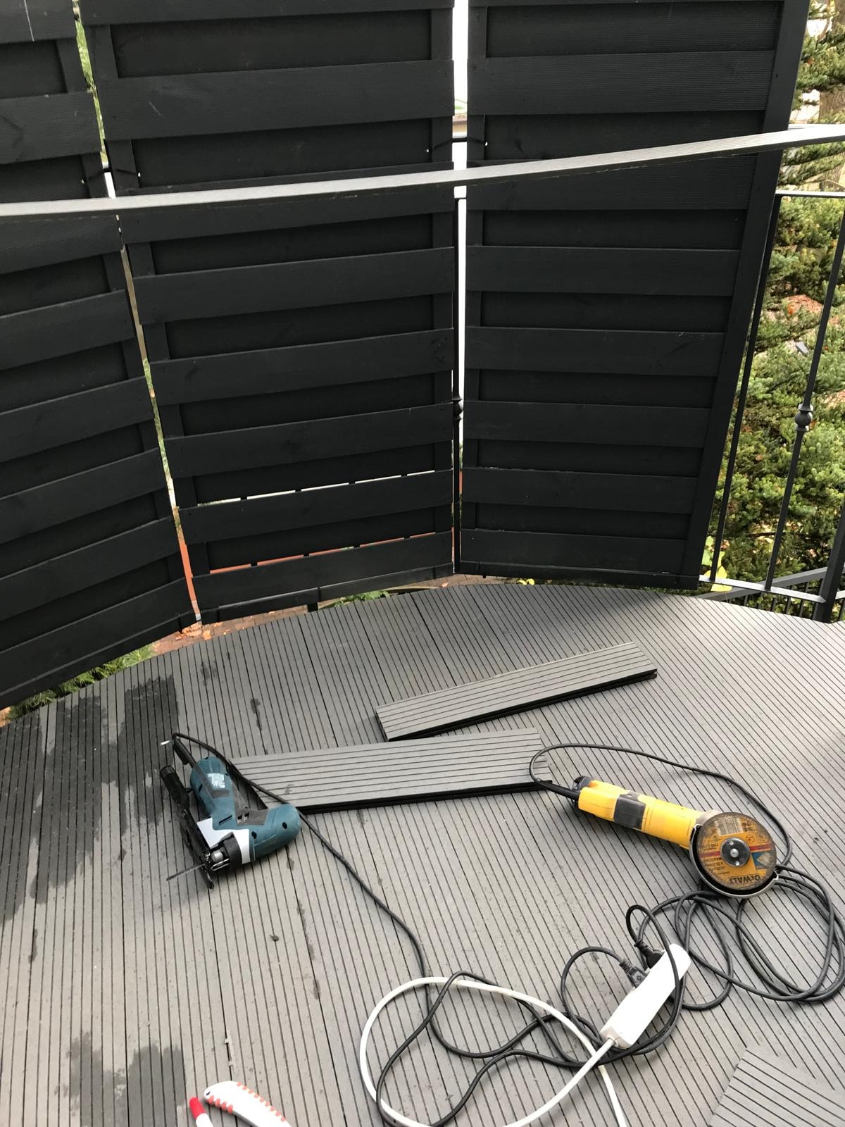 Montaż podłogi na balkonie: deski kompozytowe, wyrzynarka i szlifierka kątowa na okrągłej powierzchni balkonu z czarną balustradą z desek.