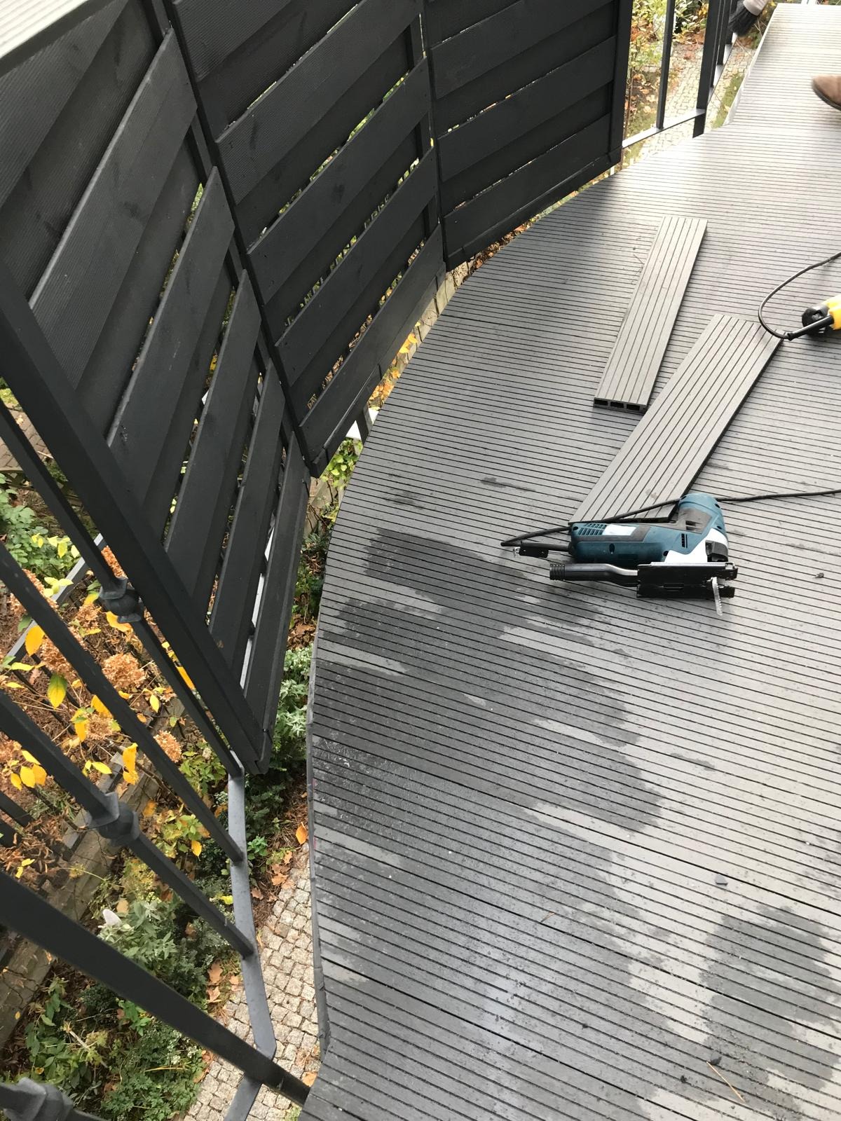 Montaż desek kompozytowych na zaokrąglonym balkonie, widoczna wyrzynarka akumulatorowa, czarna balustrada i fragment buta w prawym górnym rogu.